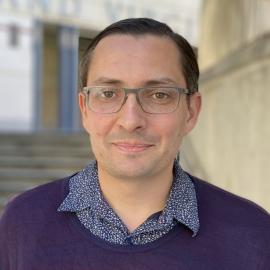 A Latino man in his mid-40s with fair skin and short, neatly side-parted hair styled to the right. He has hazel eyes, eyebrows matching his hair color, and a clean-shaven beard. He wears rectangular metal-framed eyeglasses and a blue button-up shirt with white accents layered under a bluish-purple sweater with burgundy stripes. He is facing forward against a blurred background showing a brownish-gray staircase with a railing, and at the top, a property with white walls.